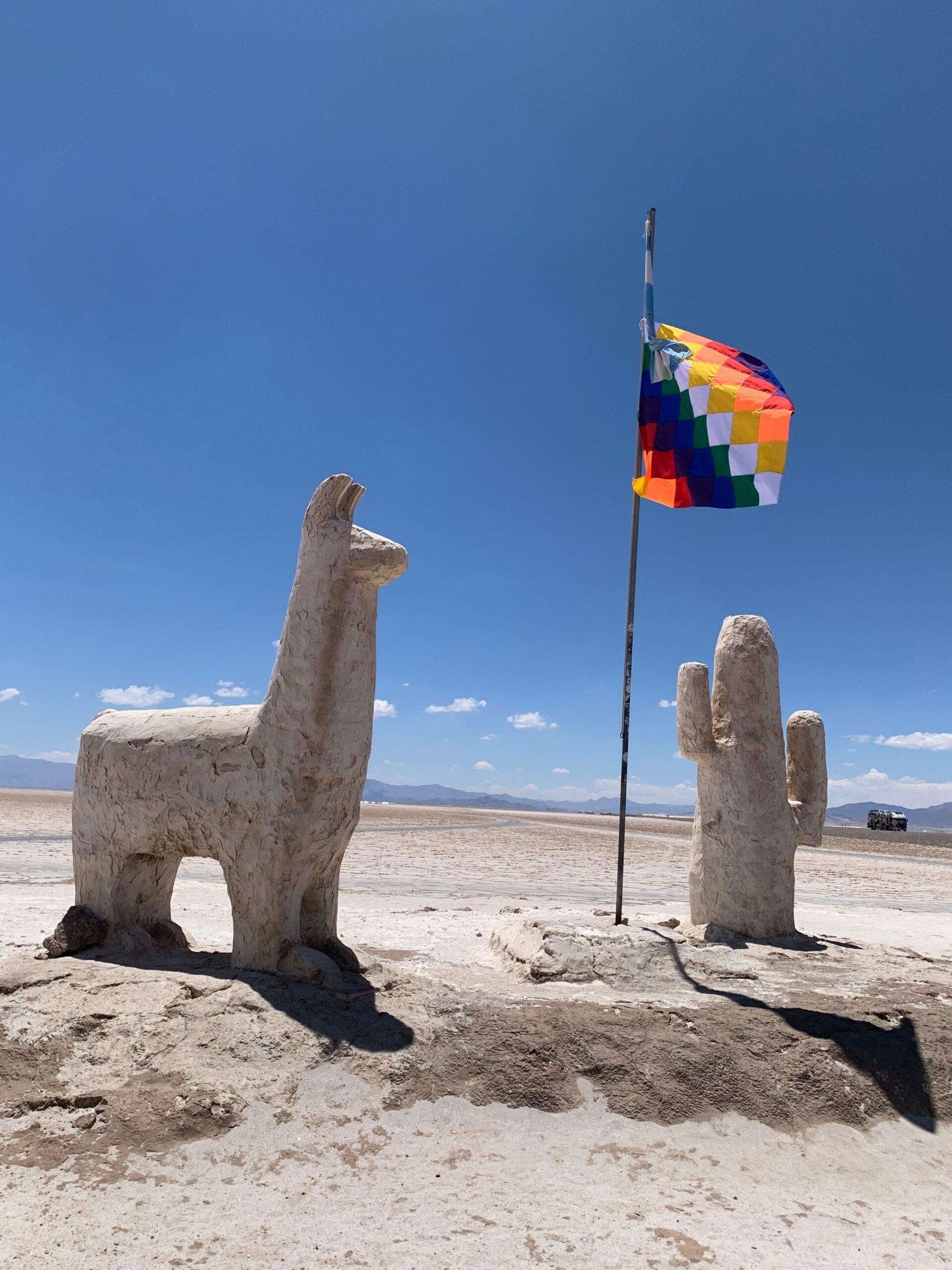 Stone llama and cactus sculptures at Salinas Grandes with colorful flag under blue sky.